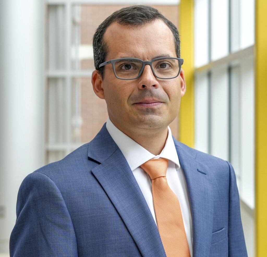 Bruno Pedreira professional headshot. Pedreira is posed in front of windows and is wearing a blue suit with an orange tie.
