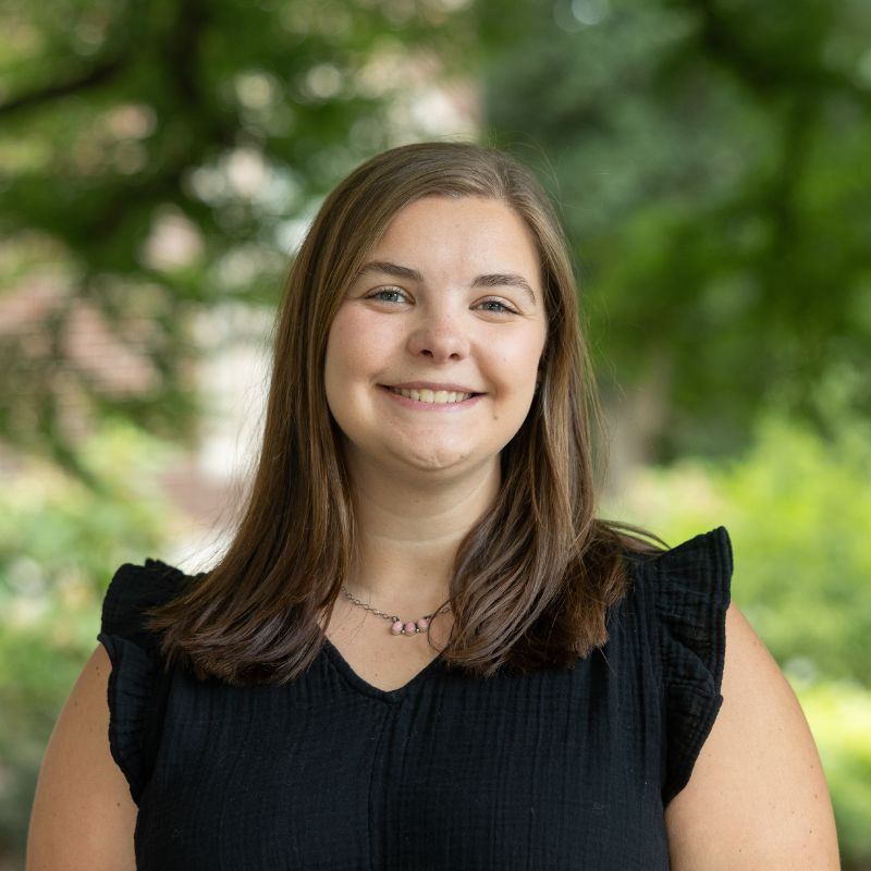 Malerie Fancher's official headshot. Fancher is posed in front of green trees and is wearing a black shirt.