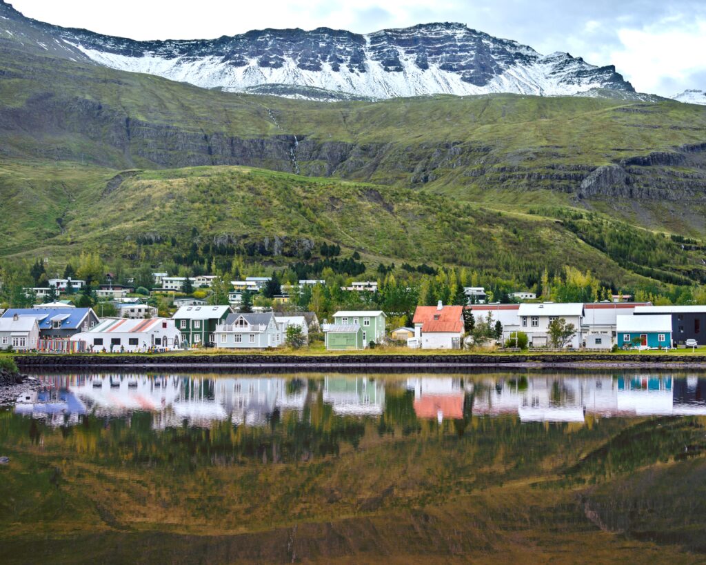 A line of buildings across a lake in front of green, snow-topped mountains.