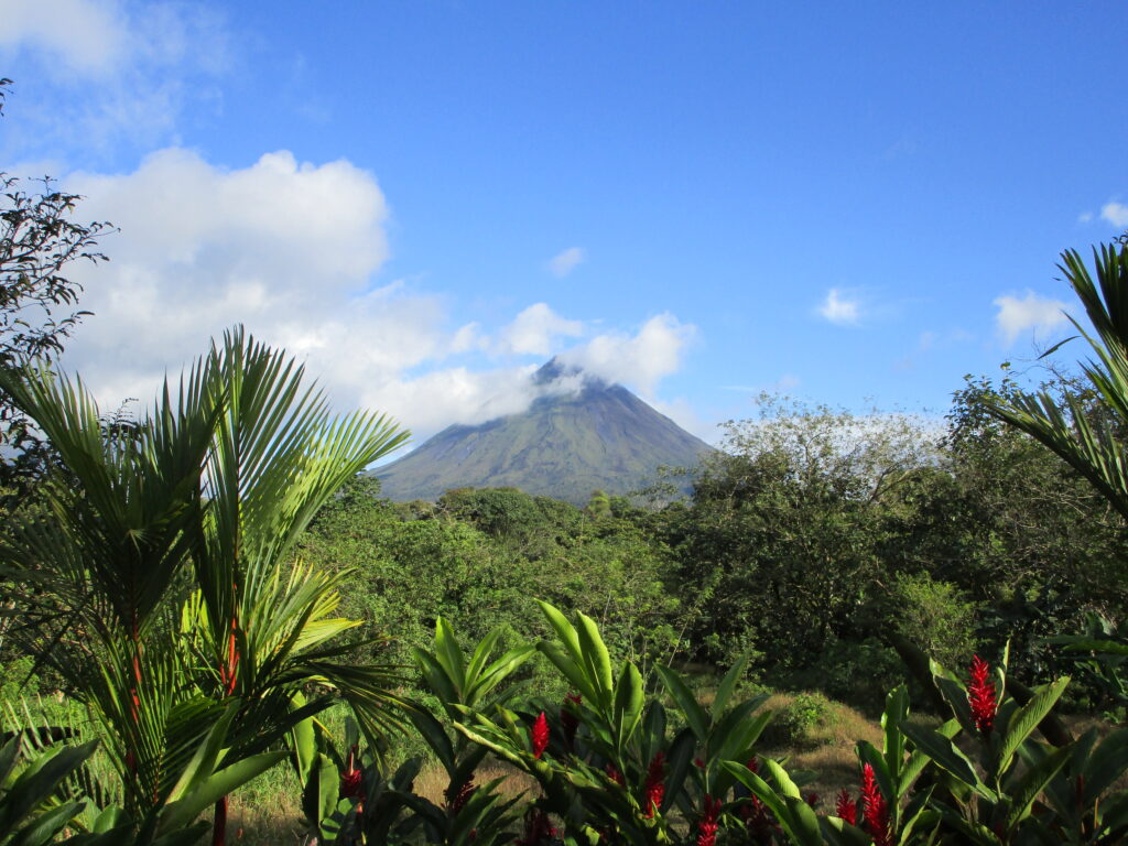 The Arenal Volcano on a sunny day. A few clouds are in the sky; the volcano is surrounded by bright greenery.